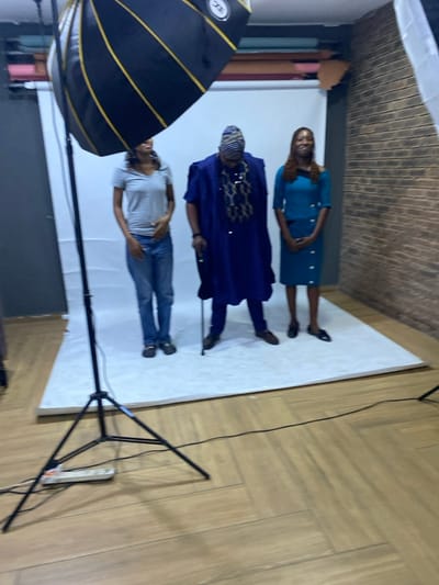 The image captures an indoor photography studio scene, likely in Animashawun, Nigeria, with three individuals posing on a white seamless backdrop. The backdrop extends from the floor up the wall, while the visible floor outside the white area is light-toned with a herringbone pattern, and the background wall displays a brick pattern.
From left to right, the first person is a man wearing a light grey t-shirt, blue jeans, and glasses resting on his head. He stands in a relaxed pose with his hands in his pockets. The middle individual, a man, is dressed in a royal blue traditional African garment, possibly an agbada, featuring gold or light-colored embroidery on the chest and a striped cap (fila). The third person, a woman, wears a teal or turquoise dress with a matching jacket, her hands clasped in front of her.
Photography equipment is visible around the subjects. A large black softbox with golden interior accents and a small circular "ECC" logo is positioned on a stand in the bottom left foreground. On the right, another black light stand with a tripod base is present, from which a black electrical cord runs across the patterned floor to a white power strip. Rolls of various colored backdrop papers can be faintly seen stacked in the far left background, confirming the studio setting. The scene depicts a photoshoot in progress.