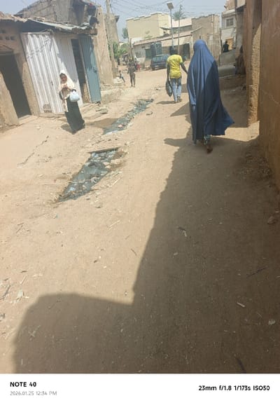 An unpaved, dusty street in Gwale, Nigeria, is depicted under bright sunlight. The street is flanked by single-story buildings constructed from mud or unpainted plaster, with visible signs of weathering. A narrow, shallow drainage ditch runs down the center-left, containing dark, stagnant water and an accumulation of plastic waste and general refuse.
In the mid-ground, a woman wearing a light-colored head covering and a dark dress walks towards the left side of the frame, carrying a light blue bag on her left arm, with her head turned slightly towards the camera. Ahead of her, a male figure in a bright yellow short-sleeved shirt and blue trousers walks away from the camera, carrying a dark bag over his left shoulder. Further ahead and to the right, a person clad in a dark blue, full-length garment (possibly an abaya or burqa) walks away from the camera, revealing patterned trousers at the ankles. Several indistinct figures, including children, are visible further down the street.
The buildings in the foreground on the left feature a corrugated metal door, partially open, and a closed blue door. In the background, more varied structures are visible, some appearing multi-story with lighter colored facades. A dark-colored car is partially visible in the distance. A solar-powered streetlight on a pole stands in the background, along with utility poles and overhead wires. Long shadows from people and buildings are cast across the dusty ground, indicating a sunny day. The camera's shadow is visible in the bottom-left corner of the frame.