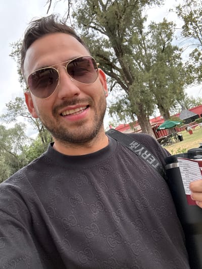 A man with sunglasses and a beard smiles. He wears a patterned black shirt and a strap that says "TERYX." A black cylindrical object is near his shoulder. Trees and a red building are visible in the background. The sky is overcast. The image is taken from a low angle.
