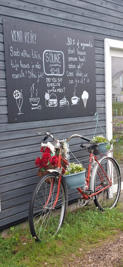 A bright red bicycle, decorated with red and white flowers in the front basket and small white flowers in hanging pots on the handlebars and seat, is parked on a gravel path in front of a dark gray wooden wall. A blackboard sign is mounted on the wall behind the bicycle. The sign is handwritten in white chalk and contains text and illustrations. 

The text on the blackboard includes phrases in Estonian: "KENA KEIK!" (Nice ride!), "Saab kõhu täis, tuju heaks ja näo naerule! Tõsi jutt!" (You can fill your stomach, improve your mood, and bring a smile to your face! True story!), and "SÕUKE Suvekohvik 10-18" (SOUKE Summer Cafe 10-18). To the right of the Estonian text, there is an English quote: ".80% of arguments start because someone hasn't eaten yet! Come inside, we can fix it!" Below the quote are illustrations of a drink, a bowl of soup, a hamburger, a slice of cake, a cup of coffee, and an ice cream cone. 

The environment is outdoors, with green grass and small yellow flowers growing around the base of the bicycle and the wall. The scene appears to be at a summer cafe in Sääre, Estonia, given the mention of "Suvekohvik" (Summer Cafe). The lighting suggests it is daytime. There are no people visible in the image. The bicycle is the central subject, adorned with floral decorations, and placed in front of a sign that advertises food and drink offerings from the cafe.