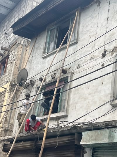 In the city of Kargil, India, a weathered, multi-story building displays signs of disrepair, with cracks and peeling plaster visible on its facade. Bamboo poles are propped against the exterior, likely serving as temporary supports or scaffolding. Several dark electrical wires crisscross the scene in front of the building. On the upper floor, two individuals are visible through an open window. One person is seated inside the window, looking downwards. Another person stands on a ledge outside the building, facing away from the camera. On a lower ledge, a third individual is crouched, appearing to work on something. This third person is wearing red pants and a light-colored shirt. The scene suggests ongoing or recent construction or repair work on the building, possibly in a densely populated urban area, given the visible electrical infrastructure.