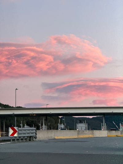 The image, when viewed in its correct orientation (rotated 90 degrees counter-clockwise), captures a dramatic sky reflected on a large, vertical surface, with highway infrastructure in the foreground.

The upper portion of the image is dominated by a vast expanse of sky, filled with dynamic, voluminous clouds. These clouds exhibit striking reddish-orange and pink hues, suggesting either a vivid sunrise or sunset. The colors are rich and warm, with darker, more shadowed areas within the cloud formations and lighter, more ethereal patches where the light is catching them. The overall sky background transitions from a lighter, pale blue-grey at the top left to a slightly darker shade around the cloud masses.

To the left and slightly below the sky, a large, rectangular, reflective panel or surface stands prominently. This surface acts like a mirror, clearly displaying a reflection of the vibrant, reddish-orange clouds from the sky above. The reflection is clear enough to show the shapes and colors of the clouds, although slightly more diffused than the actual sky. The right edge of this reflective panel features a light-colored, possibly white or off-white, frame or support structure, with visible fasteners or bolts running along its length. Several small, dark, protruding objects, possibly light fixtures or sensors, are attached to or near this panel.

The lower right portion of the image reveals a section of what appears to be a highway or a large road. The road surface is dark grey asphalt, stretching horizontally across the frame. Adjacent to the road, on its left side, is a concrete barrier or median, topped with a bright yellow painted line. Further to the left, a gap or open space suggests an underpass or a break in structures. In the mid-ground, beyond this gap, dark green, undulating hills or mountains are visible, indicating a natural landscape in the distance.

Various elements of highway infrastructure are present in this area. These include several small, light-colored structures that resemble toll booths or small roadside buildings, along with multiple poles and horizontal barriers. Closer to the foreground, at the bottom right, a prominent red and white chevron sign is visible, indicating a change in direction or a hazard. This square sign features a red arrow pointing to the right on a white background, mounted on a metal support structure. Other metal frameworks and gantries can be seen extending over parts of the road area.

The overall impression is one of a striking natural phenomenon (sunset/sunrise clouds) juxtaposed with man-made infrastructure, with the large reflective panel creating an intriguing visual doubling of the sky. No discernible text is visible on any signs or structures.