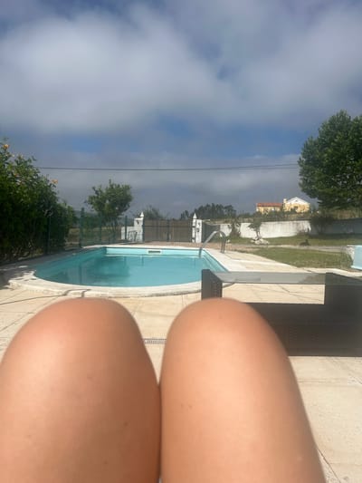 This is a first-person perspective image taken from someone relaxing by a swimming pool. The foreground is dominated by the person's legs, out of focus. In the midground is a rectangular swimming pool with clear blue water and a metal ladder. The pool is surrounded by light-colored tiled paving. A dark-colored outdoor furniture set, possibly a sofa or chairs, is visible to the right. Beyond the pool, there is a wooden fence and greenery. In the background, a house with an orange roof is visible, and trees and power lines are present against a cloudy sky. The overall setting appears to be a residential property with outdoor leisure facilities. The location is identified as Caldas da Rainha, Portugal.