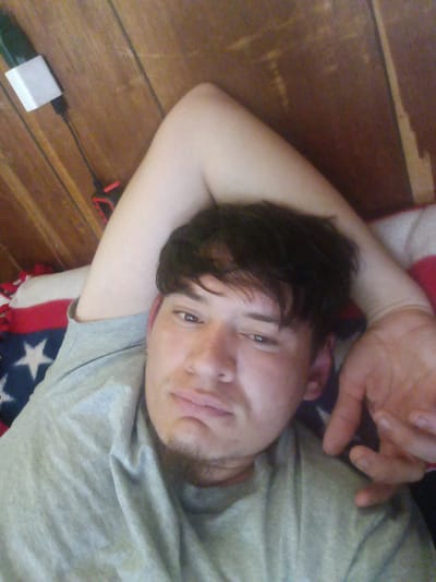 A person is lying down on a bed with a blanket featuring an American flag design. The person is fair-skinned, with dark hair and stubble, wearing a grey t-shirt. Their right arm is bent over their head. The background consists of wooden paneling. A white charger is plugged into an outlet near the head of the bed. The location is unknown but is hypothesized to be in the United States. The city is unknown but is hypothesized to be Park Hill.