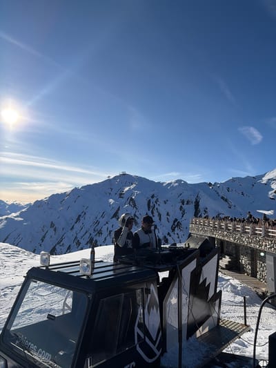 The image depicts an outdoor DJ event on a snow-covered mountain environment near Schwendau, Austria, specifically at the Schneekarhütte. In the foreground, two male individuals are positioned behind a black Pioneer DJ console, which is mounted on the rear platform of a black tracked vehicle, likely a snow groomer. The individual on the left wears a white helmet and headphones, while the individual on the right wears a dark cap, sunglasses, and headphones. A bottle rests on the roof of the vehicle's cabin, which displays text including "schneekarhutte.at". Large black speakers are visible next to the DJ setup. To the right, a multi-level building constructed of stone and wood features a prominent wooden balcony or terrace. Approximately 30-40 individuals are gathered on this balcony, observing the scene or viewing the landscape. The background features extensive snow-covered mountain ranges under a clear, bright blue sky with sparse white clouds. The sun is visible in the upper left, casting a strong glare. A ski lift station is visible on a distant mountain peak.