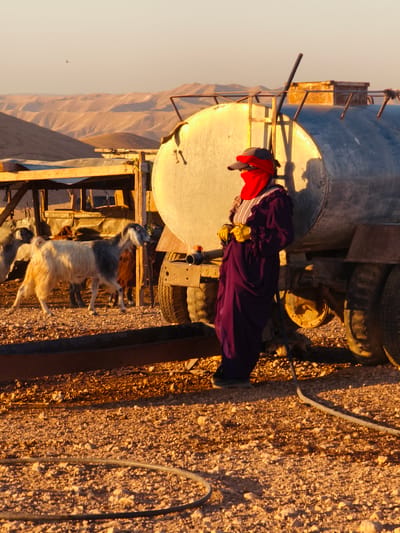 In the city of Keidar, Palestinian Territories, a person wearing a dark purple robe, a red face covering, a dark cap, and yellow gloves stands next to a large, circular metal water tank on a truck. The person is positioned in front of the tank, with their body angled towards the viewer. A dark hose is visible on the ground in the foreground, extending from the bottom left of the frame towards the middle right. Several goats are present in the background, standing near a wooden structure with a corrugated metal roof. The environment appears arid, with dusty, rocky ground and rolling hills in the distance under a pale sky. The lighting suggests either dawn or dusk, with warm tones illuminating the scene.