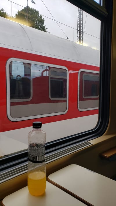 A slightly angled, medium shot captures a view from inside a train carriage, looking out a window at another train. In the foreground, a plastic bottle filled with a yellow liquid, likely juice, stands on a folded-down tray table. The bottle has a black cap and a label with text and images. The tray table is light beige with a wooden trim. The window frame is dark, and the outside is dominated by the exterior of a red and white train. The red section has several rectangular windows, some of which reflect the interior of the train. Beyond the train, an overcast sky is visible, along with trees and a metallic pylon with overhead wires. The lighting is diffused, suggesting an outdoor scene on a cloudy day. The composition creates a sense of travel and contemplation. The location is the train station in Vrútky, Slovakia.