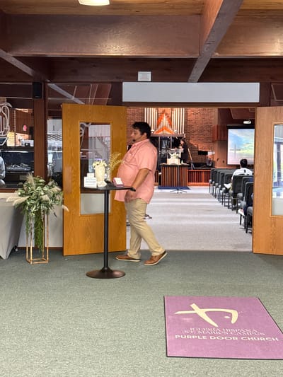The image shows the interior of a church, identified by a sign as "Iglesia Hispana St. Mark's Campus Purple Door Church" which indicates it is in Columbus, United States of America. A man in a pink shirt and tan pants stands behind a wooden podium with floral arrangements and a guest book. The podium is in front of a wooden-framed window with a view of a church service in progress, with attendees seated in rows of chairs facing a stage with musical instruments. A purple rug with the church name is on the floor.