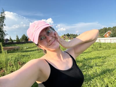 A woman wearing a pink baseball cap and black tank top takes a selfie outdoors in a grassy area. She is smiling and touching her face with her hand. In the background, there are trees, a fence, and a few houses, suggesting a rural or suburban setting. The sky is blue with white clouds.