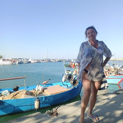 A medium shot captures a woman standing on a concrete dock in the harbor of Limani Litochorou, Greece. The woman is positioned to the right of the frame, facing towards the viewer with her right hand on her hip. She is wearing a white, sheer cover-up with a blue coral-like pattern, which is open to reveal dark swimwear underneath. She has sunglasses perched on her head and her hair is pulled up. Her legs are bare, and she is wearing blue and white flip-flops. To the left of the woman is a blue fishing boat with various pieces of equipment, including nets and buoys, on board. The boat is partially moored to the dock. In the background, several other fishing boats and sailboats are visible in the calm blue water of the harbor. A clear blue sky with no clouds forms the upper part of the image. The sunlight casts distinct shadows on the concrete dock from the woman and the boats. The overall scene depicts a sunny day at a Greek harbor with a woman posing by a fishing boat.