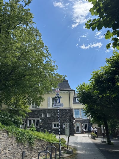 The image captures a sunny day scene in Königswinter, Germany, outside a building that appears to be a restaurant or Biergarten. The building itself has a lower level constructed of dark stone and an upper level painted yellow with white shutters. A sign with blue and white stripes and the word "Biergarten" is prominently displayed near the entrance. Green trees with lush foliage frame the scene, with the blue sky and scattered white clouds visible above. A paved walkway leads to the building's entrance, and several people are visible near the entrance, some standing and one walking. The environment suggests a relaxed, outdoor setting typical of a German Biergarten.