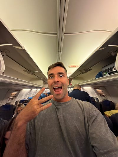 The image shows a man in the foreground with a surprised expression, taking a selfie inside an airplane. He is wearing a grey t-shirt and making a peace sign with his right hand. Behind him, the interior of the airplane is visible, with rows of seats and overhead compartments. Other passengers can be seen in the background, some with their heads down. There is a red sign with white text that says "KIJÁRAT EXIT" in Hungarian, indicating an exit. The lighting suggests it is either daytime or the cabin lights are on. The scene is consistent with being on a commercial flight.
