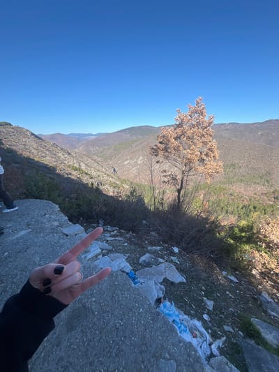The image displays a mountainous landscape near Tuhovishta, Bulgaria, under a clear blue sky. In the immediate foreground, a left human hand, adorned with black nail polish and a silver ring on the ring finger, performs a gesture where the index and pinky fingers are extended upwards. This hand emerges from a dark long-sleeved garment. The hand is positioned above a grey, rough-textured concrete ledge that occupies the lower-left section of the frame. Below and to the right of this concrete structure, the ground slopes steeply downwards, consisting of dirt and small rocks, with visible litter including blue and white plastic materials. A deciduous tree with light brown, dried leaves stands prominently on this slope towards the right of the mid-ground. Bare bushes and sparse, wintry vegetation cover the hillsides in the mid-distance. To the left mid-background, the lower legs and feet of a second individual, wearing light-colored footwear and dark pants, are partially visible. The background reveals a deep valley flanked by multiple layers of mountains and hills, predominantly covered in dormant vegetation and some patches of evergreen trees. The sky is uniformly clear and bright blue, indicating sunny weather.
