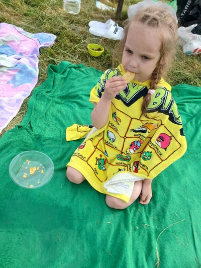 In this overhead shot, a young girl sits on a green towel outdoors, wearing a bright yellow poncho featuring Angry Birds characters. She is holding a cracker to her mouth, with a small plastic container holding remnants of crackers next to her. To her left, white slides and a yellow bowl are visible on the grass. A light-colored towel with a tie-dye pattern is spread out on the ground to her lower left. The scene appears to be a casual outdoor gathering, possibly a picnic or a day at the park, given the grassy setting and relaxed atmosphere. The lighting suggests daytime. The overall impression is one of a candid, everyday moment captured during an outdoor activity.