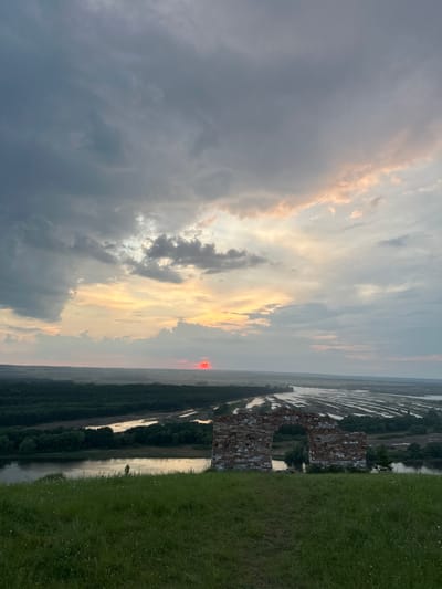 The image depicts a scenic view of the Volga River delta near Nizhniye Vyazovye, Russia, during sunset. The foreground is dominated by a grassy hill with scattered wildflowers and a decaying brick archway, possibly a remnant of a historical structure. In the middle ground, the wide Volga River meanders through a lush green landscape, reflecting the soft hues of the sky. The background reveals a vast expanse of agricultural fields, possibly flooded for rice cultivation, creating a patterned effect. The sky is filled with dramatic clouds, painted with a blend of grays, whites, and warm oranges and pinks as the sun, a vibrant red orb, dips below the horizon. The overall atmosphere is tranquil and picturesque, capturing the beauty of the Russian countryside at dusk.