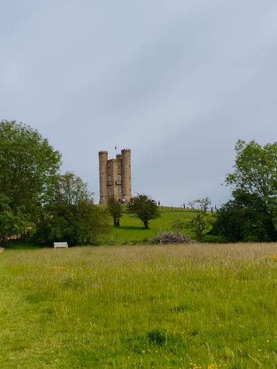 This image depicts Broadway Tower, a striking landmark situated on a hill in the Cotswold District of the United Kingdom. The tower, made of light-colored stone, features multiple levels with arched windows and crenellated battlements, and a flag flies from its peak. Several people are visible near the base of the tower, suggesting it's a tourist attraction.
The scene is set amidst rolling green hills and lush meadows. A white bench sits in a field of tall grass in the foreground, inviting viewers to imagine a moment of rest. Trees frame the tower on either side, with a particularly large, dense tree on the left and a cluster of trees and bushes on the right, creating a sense of depth. In the middle ground, a pile of weathered branches rests on the grass.
The sky is overcast with a pale, diffused light, indicating it is likely daytime, possibly late morning or afternoon. The weather appears calm and temperate, suitable for outdoor activities. The overall atmosphere is serene and picturesque, capturing the natural beauty of the Cotswolds. There is no visible text in the image.