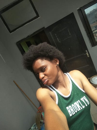 The image displays a young adult female, with dark complexion and short, dark, afro-textured hair, captured in a self-portrait in Cotonou, Benin. She wears a green tank top with white trim, featuring "BROOKLYN" in white lettering across the chest; a small dark stud earring is visible in her left ear. Her lips are slightly pursed, and she looks directly at the camera, her right arm extending into the foreground. The background, illuminated by strong natural daylight, reveals a dark gray wall on the left against which a wooden stick or broom handle leans, with a pile of miscellaneous items including plastic bottles and broom bristles at its base. To the right of the subject, a dark brown door with a light-colored handle/lock mechanism is partially visible, adjacent to a window offering a blurred view of distant city buildings. A white, lidded container is present behind the subject's right shoulder. An overhead light gray wall section contains a dark-framed rectangular opening.