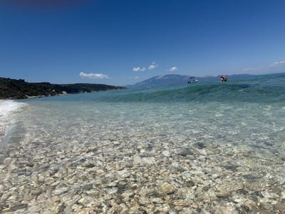The image shows a crystal-clear shallow sea with a rocky seabed visible through the transparent water. The water gently laps at the shore, creating a soft wave on the left side of the frame. In the background, a lush green hillside slopes down towards the sea. Further in the distance, mountains are visible under a bright blue sky with a few scattered white clouds. A small white boat with an awning is seen on the water, and a person is visible swimming or floating in the sea near the boat. The scene is a tranquil coastal landscape, likely in Elaties Municipal Unit, Greece, given the visual characteristics of the vegetation, coastline, and mountainous background.