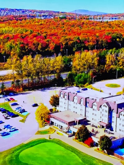 This aerial image showcases a vibrant autumn landscape adjacent to a modern building complex. The scene is dominated by a dense forest displaying a striking palette of fall colors, ranging from deep reds and oranges to bright yellows and greens. The trees are the primary natural subject, with their foliage creating a rich tapestry.

To the right of the forest, a multi-story building with a light-colored facade and reddish-brown roofs is visible. The building has numerous windows, indicating it is likely a residential or commercial structure. A parking lot is situated in front of the building, filled with several cars, predominantly in shades of blue and white. The parking lot is bordered by a bright green grassy area, suggesting a well-maintained outdoor space.

In the background, to the left of the forest, a series of tall, white structures, possibly part of a construction site or a large industrial facility, are present. A mountain or large hill is also discernible in the distance. A road or pathway winds through the trees, and a few streetlights are visible along its edge.

The image appears to be taken during the daytime, under clear weather conditions, given the bright lighting and distinct shadows. The location is identified as Gorzow, Poland. There is no discernible text within the image. The overall impression is one of a harmonious blend between nature's seasonal beauty and modern infrastructure.