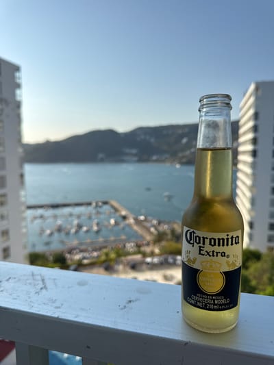 The image shows a bottle of Coronita Extra beer lying on a white painted ledge. The bottle is partially full, with condensation visible on its surface. In the background, a blurred view reveals a bay filled with boats, bordered by a hillside and buildings. The location is likely Puerto Marqués, Mexico, based on the brand's association with Mexico and the harbor scene. The sky is a clear, pale blue. The photograph is taken at eye-level, focusing primarily on the beer bottle.