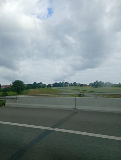 A wide-angle, eye-level shot captures a rural landscape in Wysoka, Poland, under a cloudy sky. The foreground features a paved road with a white line marking its lane, suggesting it's part of a highway or major road. A light-colored concrete barrier runs along the side of the road, separating it from the surrounding environment. Beyond the barrier, rolling green fields stretch into the distance, dotted with trees and agricultural fields. Several buildings, appearing to be houses or farmhouses with red-tiled roofs, are visible nestled amongst the trees. The overall scene depicts a serene, rural setting on what appears to be a mild day with overcast skies.