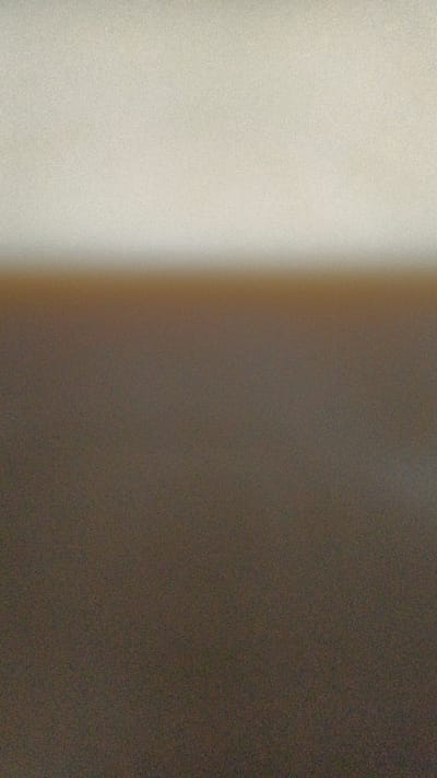 This is an abstract image with a subtle gradient. The top portion is a very light, desaturated beige, with a slight yellowish tint. This transitions into a darker, brownish-grey hue that occupies the majority of the image. The transition between the two colors is soft and blurred, creating a horizon-like effect. The entire image is textured with a fine grain, giving it a slightly vintage or artistic feel. There are no discernible objects, people, or specific locations visible. It appears to be a very minimalist or abstract composition.