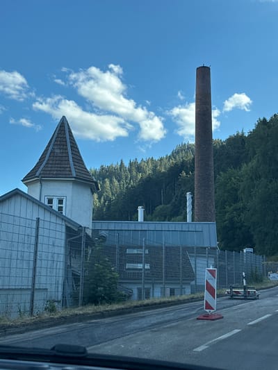 The image shows an industrial building complex with a tall, brick chimney in the background, located in Hornberg, Germany. The site is surrounded by a metal fence and situated at the base of a densely wooded hillside. A paved road with white lane markings runs in the foreground, with a temporary traffic barrier and a red and white striped pole indicating ongoing road work or a detour. The sky is blue with scattered white clouds. The architecture of the building includes a distinctive octagonal tower with a conical tiled roof and a white-paneled structure.