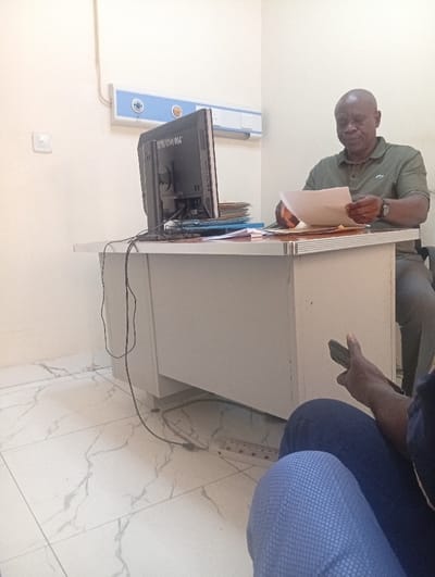 An indoor scene in Okengwe - Obehira, Nigeria, depicts a room with light-colored walls and white ceramic floor tiles patterned with grey veining. A bald, dark-skinned male, wearing an olive green short-sleeved polo shirt and a watch on his left wrist, is seated behind a light-colored desk. He is actively examining multiple sheets of white paper held in both hands. To his left on the desk is a black flat-panel computer monitor, viewed from the rear, with cables extending towards the floor. Stacks of folders and papers are present on the desk. Mounted on the wall above the desk is a white and blue horizontal panel featuring two circular ports with blue rings and several white rectangular switches or outlets. A white wall switch or outlet is also visible on the left wall. In the foreground, the lower body of a second individual, dressed in dark blue trousers, is visible, with a right hand holding a black smartphone. A white multi-socket power strip with multiple plugs inserted lies on the floor beneath the desk, connected by black cables.