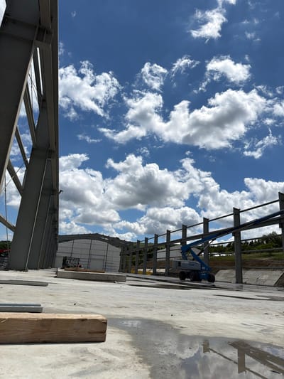 An outdoor, low-angle, vertical-format shot captures a construction site under a partly cloudy blue sky. In the foreground, a rough wooden beam rests on a wet, concrete surface. In the midground, a blue Genie lift, with the model number "S-85" visible, is positioned adjacent to a series of unfinished steel beams. Beyond the lift, a large, white building with a curved roof is under construction. The background features a treeline and a rolling hill. The overall scene depicts an active construction zone.