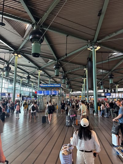The image depicts a bustling scene inside the Schiphol Railway Station in the city of Schiphol, Netherlands. The station is populated with numerous people, many pulling luggage, suggesting they are travelers. The focus is on the interior architecture, characterized by a high, green-painted, steel-framed ceiling with integrated lighting and dark ventilation or speaker systems. Signage indicating "Schiphol Railway Station" is visible, along with train departure and arrival information displays. The floor has parallel lines that run into the distance, contributing to the sense of movement and direction in the station. The general impression is one of a busy transportation hub.