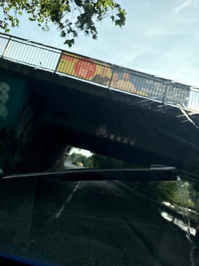 This media file shows a view looking upwards at a bridge structure against a bright, overcast sky. Green foliage from trees is visible on the left side of the frame, suggesting an outdoor, daytime setting. The bridge itself is dark and appears to be made of metal and concrete. A section of the bridge has a colorful banner or advertisement attached, featuring yellow and red elements with some text. Some of this text is partially obscured and reversed due to reflection.

On the right side of the image, a highly reflective surface, likely a window or a polished exterior, mirrors the surroundings. This reflection shows blurred green foliage and hints of light, suggesting the presence of trees and possibly buildings or structures nearby. The reflection is distorted and somewhat abstract due to the curvature or imperfections of the surface.

The overall impression is of a slightly abstract, urban environment. The lighting suggests it is daytime, possibly overcast given the bright but diffused sky. There are no clear indications of specific events or emotions, but the upward perspective and the presence of urban infrastructure hint at a scene observed from below or possibly from inside a vehicle. The reflected text on the banner is too unclear to be definitively read, but it appears to be in a Latin-based alphabet.