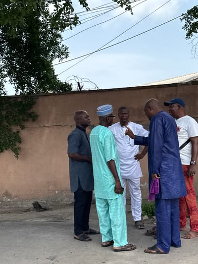 A ground-level, medium shot depicts five men standing in a line outdoors, likely in Falomo, Nigeria. The man closest to the camera wears a dark gray shirt and black pants. Behind him is a man in a light blue outfit with a blue and white striped hat and glasses. Next in line is a man dressed in white, with glasses. The fourth man wears a blue outfit and holds a purple object in his hand. The man furthest from the camera has on a white t-shirt with a black strap and red and orange pants. All men wear sandals. The background consists of a weathered, light-brown wall with barbed wire at the top and green vines growing on it, along with a gray sidewalk and a tree with dense foliage. Overhead power lines are visible against the light blue sky.