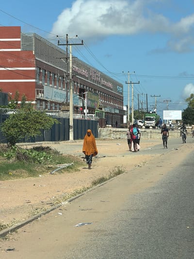 The image shows a street scene in what is likely the city of Muxara, Mozambique. A building with "Centro Medico" signage and a red brick base is visible, with utility poles and lines running along the street. People are walking on the roadside, including one individual wearing a brown garment covering their head and body. A truck and other pedestrians can be seen further down the road. The road is asphalt with a dirt shoulder, and the sky is blue with scattered clouds.