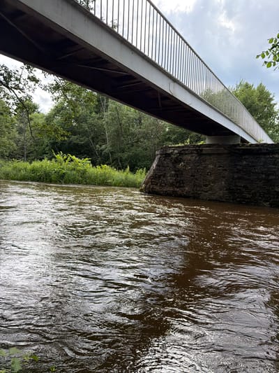 The image shows a pedestrian bridge spanning a muddy, flowing river. The bridge has a light-colored metal railing and a darker metal underside. The riverbanks are lined with lush green vegetation and trees. A stone support structure anchors the bridge on the right side of the image. The sky is overcast with clouds. The location is identified as Tēraudi, Latvia. There are no people visible in the image.