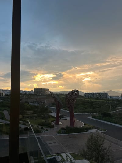 The image shows a view of the city of Zibatá, Mexico, at dusk. The sky is filled with dramatic clouds, with a vibrant band of sunlight breaking through on the horizon, illuminating the landscape. In the foreground, a large, dark vertical beam, likely part of a building structure, frames the scene on the left.
Dominating the mid-ground are two large, abstract sculptures resembling stylized trees with intricate, openwork designs. They are colored in a muted red. Surrounding the sculptures is a landscaped area with green grass, pathways, and scattered rocks.
Further in the background, a circular, multi-story building with visible construction scaffolding is situated. To its right, a modern, multi-story residential building with numerous windows is visible. Beyond these structures, a chain of mountains forms the backdrop against the dusky sky. The overall environment appears to be a planned community with modern architecture and green spaces. There are no people visible in the image. The lighting suggests either sunrise or sunset, with the golden hues of the sun reflecting off the clouds and casting a warm glow over the scene.