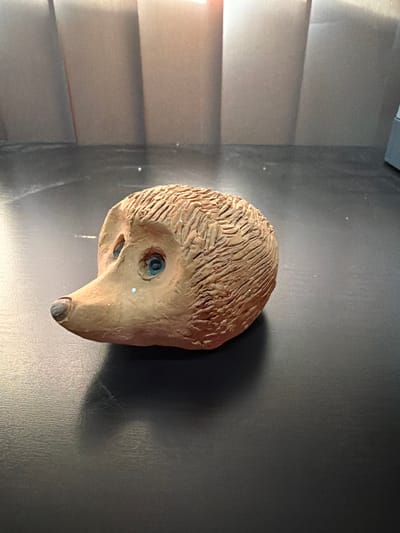 The image shows a ceramic hedgehog figurine placed on a dark, textured surface. The figurine is light brown with carved lines to represent fur and has two blue eyes and a darker brown nose. A light is reflecting off of the surface and also onto the hedgehog. The background consists of vertical, ribbed panels in muted tones. The scene is indoors, likely on a table or countertop. There is no indication of Bissendorf, Germany in the image.