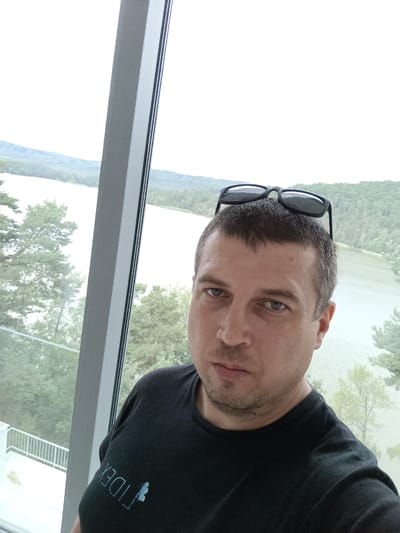 The image is a selfie taken by a man with short, brown hair and a light beard. He is wearing a black t-shirt with a blue logo that appears to read "FIDEX" with a small blue butterfly or insect graphic. Sunglasses are resting on top of his head. He is positioned in front of a large window, which offers a view of a body of water, possibly Lake Mácha, surrounded by green hills and trees. The window frame is a prominent vertical element on the left side of the image. The lighting suggests it is daytime, possibly overcast, given the soft, diffused light. The overall impression is of a casual, outdoor or semi-outdoor setting, possibly a modern building overlooking the lake.