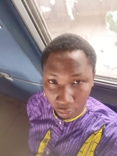 A young man with short, dark hair is wearing a purple and yellow striped shirt. He has dark eyes and a serious expression. He appears to be indoors, with a window visible in the background. The window has a metallic frame. The lighting suggests a bright day outside. He seems to be taking a selfie or posing for a photo.