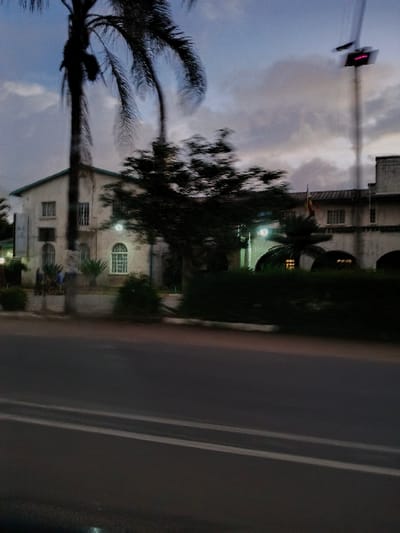 This image depicts an outdoor street scene in Marondera, Zimbabwe, captured during dusk or early evening. The perspective suggests it was taken from a moving vehicle, evidenced by a slight motion blur.

In the foreground, a dark asphalt road is visible, featuring clear white lane markings – one solid and one dashed. To the left of the road, the scene transitions into a built-up area interspersed with vegetation.

Several buildings are visible, most notably a light-colored structure, possibly white or cream, with a dark grey roof. This building features multiple windows, including one with a distinct arched top and white frames. Artificial lights are illuminated around the building, suggesting the fading daylight. Further along, another building section with rectangular windows can be seen.

The landscape is rich with plant life, including several dark, leafy trees and prominent palm trees. A large palm tree is silhouetted against the twilight sky on the far left, its fronds clearly visible. Other green foliage and bushes are scattered near the base of the buildings. High above, a traffic light displaying a red signal is noticeable on the far top-left. A small flag, appearing to be yellow and red, is faintly visible on a pole near one of the buildings. The sky is a cloudy blue-grey, typical of twilight.

No people are visible, and there is no discernible text in the image.