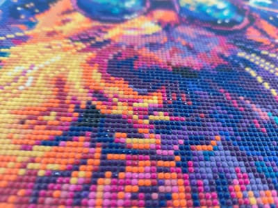 The image is a close-up, high-angle, shallow depth of field photograph of a diamond painting. The painting is composed of numerous small, round, colored resin beads arranged in a grid pattern to form a larger image. The colors visible include various shades of yellow, orange, pink, purple, and blue, suggesting a vibrant and possibly abstract or stylized subject matter. The texture of the beads is slightly uneven, with some appearing to catch the light. The focus is on the middle ground of the painting, with the foreground and background rendered out of focus. There are no people, animals, or identifiable landmarks visible in the image, and no specific location within Mykulychyn, Ukraine can be determined from the visual information provided. The scene depicts a craft or art project in progress or completed.