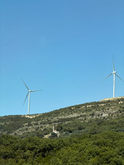 The image shows a landscape scene with two wind turbines visible on a hillside under a clear blue sky. The left turbine is closer to the viewer, with its blades extended. The right turbine is further in the distance, also with blades extended. The hillside is covered in green vegetation and some rocky terrain. In the mid-ground, nestled amongst the trees on the hillside, is a small white building with a red roof, which appears to be a chapel or a small church. The foreground is dominated by lush green trees. The scene is devoid of people. The location is Bratica - Braticë, Montenegro.