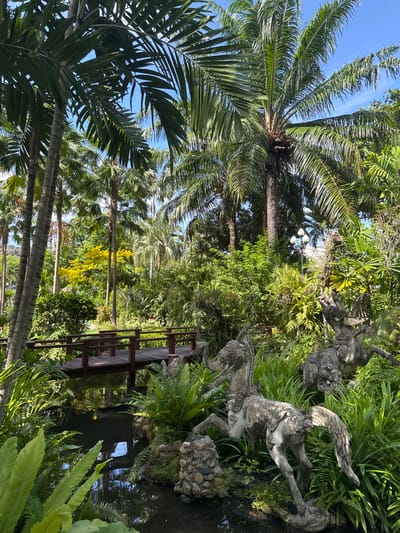 The image depicts a garden landscape in Samrong Nuea Subdistrict Municipality, Thailand, featuring tropical vegetation including palm trees. A wooden bridge spans a body of water, and statues of mythical creatures are nestled amongst the greenery.