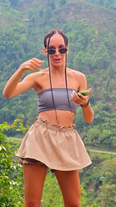 A young woman stands outdoors, captured in a full-body portrait against a lush green mountainous backdrop, indicative of Nậm Dịch Commune, Vietnam. She appears to be in her 20s or 30s, with dark hair styled in multiple long braids, some of which fall forward. Her attire includes a grey ribbed strapless bandeau top, light brown or beige high-waisted ruffled shorts that resemble a skirt, and oval-shaped sunglasses with gold frames. A delicate gold chain necklace hangs around her neck.
She is actively engaged, holding a green smartphone in her right hand while her left arm is raised with fingers slightly curled, suggesting a gesture, possibly pointing or emphasizing something. Her mouth is slightly open, conveying an impression of speech, surprise, or animated reaction. Several colorful bracelets adorn her right wrist, and her nails are manicured with a light polish.
The scene is set during daytime, with clear, natural lighting illuminating the dense foliage and rolling hills in the background. A faint road or path can be discerned winding through the distant mountains. The overall setting is a vibrant, natural landscape. No text is visible in the image.