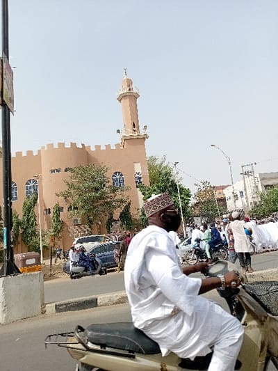 This image depicts an urban street scene in Sani Mai Nagge, Nigeria, under clear daytime conditions.
In the immediate foreground, a male subject rides a light-colored scooter, positioned in profile facing right. He wears a white traditional tunic (agbada), white trousers, a patterned kufi cap, dark sunglasses, and a black face mask, with his hands gripping the scooter's handlebars. The scooter features a black seat and a rear cargo rack.
The midground reveals a paved road with a concrete curb separating lanes. On the left, a silver sedan is visible alongside a blue scooter carrying two individuals. An orange three-wheeled rickshaw is partially obscured behind a dark vertical pole. On the right, a large gathering or procession of individuals, many dressed in white traditional garments, occupies the roadway. Several other motorcycles with riders are interspersed within this group.
The background is dominated by a tan-colored mosque, characterized by a crenellated parapet along its roofline and numerous arched windows with blue decorative grilles. A tall, slender minaret, featuring a balcony and topped with a finial (crescent moon and star), rises from the mosque. Green trees are distributed around the mosque and along the street. The sky above is clear and light blue, with additional urban buildings and utility structures visible in the far distance on the right.