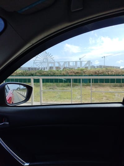 The image, taken from inside a car, shows a view through the side window and side mirror. In the distance, large white lettering is visible against a blue sky with scattered clouds. The lettering appears to spell "VINOGRADNY," identifying the location as Vinogradny, Russia. Behind the lettering, construction cranes and unfinished buildings suggest an ongoing development. A white fence runs horizontally across the middle ground, separating the road from a grassy field. In the side mirror, a red car is reflected, driving on a road. The car interior, including a black door panel and a chrome door handle, is visible in the foreground.