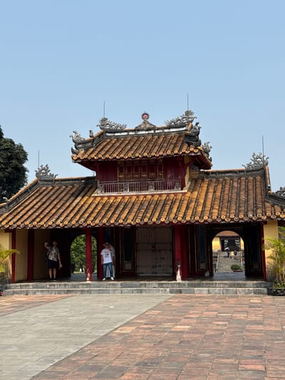 A medium-angle shot captures a building with an ornate, multi-tiered roof covered in rounded terracotta tiles. The structure is adjacent to a brick pathway. The roofline features decorative silver accents. The building has red-painted doors and columns. Two people stand underneath the structure. One of them is a man wearing a short-sleeved shirt and shorts, holding a phone. The other person is wearing a white t-shirt and jeans. In the background, through a circular door, a staircase and two people can be seen. The sky is clear and blue. The location is Kim Long Ward, Vietnam.