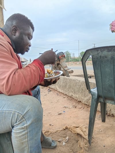 The image depicts a man sitting on a green plastic chair by the side of what appears to be a road in Akute, Nigeria. He is wearing a red shirt and blue jeans, and is eating from a plate. A motorcycle is parked nearby. The ground is dirt and sand. Overhead power lines and poles are visible in the background. The scene is captured from a low angle, slightly rotated.