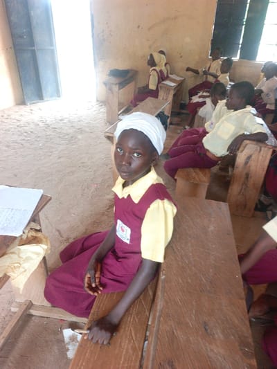 This image depicts a classroom scene in Mada, Nigeria. In the foreground, a young girl with dark skin, wearing a white head covering, a yellow collared shirt, and a maroon sleeveless tunic with a white badge, is seated on the earthen floor, looking directly at the camera. Her right hand rests on a worn wooden desk, possibly holding a writing implement. On the desk to her left, a sheet of paper with handwritten text and crumpled light-colored paper are visible.

Behind her, several other children, similarly dressed in maroon and yellow school uniforms, are seated at simple, worn wooden desks and benches. Their postures vary; some appear attentive, while others lean back or look within the room. One child in the far back left also wears a light head covering.

The classroom features rough, light brown walls and an uneven, sandy earthen floor. A brightly lit doorway or large opening on the left side of the frame provides significant natural light, while another smaller window opening is visible high on the back right wall. The wooden furniture shows signs of extensive use, with visible scratches and wear.