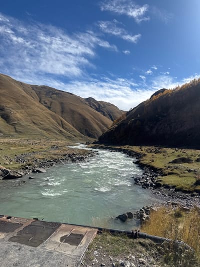 The image depicts a panoramic view of a river valley in Kvemo Okrokana, Georgia, under a partly cloudy blue sky. A milky, pale turquoise river with significant rapids flows from the distant background towards the immediate foreground, occupying the central and lower-mid sections. Its banks are characterized by dark rocks and sparse, dry grass.
To the left of the river, large, rounded mountains with slopes covered in yellowish-brown dry grass dominate the landscape, extending into the background. A small, white, rectangular structure is visible high on the left mountain slope. A dirt track or unpaved road runs parallel to the river on the left bank, flanked by several wooden utility poles with visible wires extending into the distance. The terrain adjacent to this track is relatively flat and grassy.
To the right of the river, a darker, steeper mountain slope, partially in shadow, rises sharply. The upper part of this slope is adorned with evergreen trees and deciduous trees exhibiting yellow and orange autumn foliage.
In the immediate foreground, a dark, flat, metallic or concrete structure with embossed rectangular patterns is visible on the lower left, extending partially over the river's edge. On the lower right, a dark pipe runs horizontally along the grassy bank before disappearing towards the river. The sky above is bright blue with scattered, wispy white clouds. No people or animals are visible.