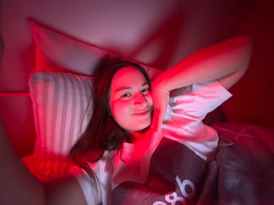 The image captures a young woman lying down, viewed from an overhead perspective, with her head towards the bottom right of the frame and her body extending towards the top left. The entire scene is bathed in a strong, reddish light, creating a warm and atmospheric ambiance.
The woman has long, dark hair that cascades around her head, resting on a striped pillow. She is wearing a simple white short-sleeved t-shirt. Her right arm is bent, with her hand gently cupping the side of her face near her ear. Her left arm is mostly out of view, possibly tucked under a blanket. She looks directly at the camera with a soft, relaxed expression, possibly a faint smile.
She is lying on what appears to be a bed. Her head rests on a pillow with alternating light and dark stripes, which itself is on top of a larger, lighter-colored pillow. A dark-colored blanket or duvet, possibly patterned, covers her upper torso and arms. On this dark fabric, positioned above her head, white letters are visible, appearing as "9b" or "qb" when read from her perspective (inverted in the current image orientation). The background is mostly dark and indistinct, suggesting an indoor setting like a bedroom, with the red lighting being a prominent feature of the environment rather than natural light.